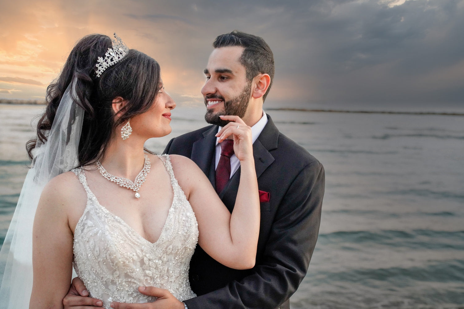 A newlywed couple shares a romantic moment by the beach in Cyprus during sunset. The bride wears a sparkling white gown with a tiara, while the groom is dressed in a black suit with a red tie. They gaze into each other's eyes, with the ocean and a dramatic sky in the background, capturing the essence of a dreamy Cyprus wedding.